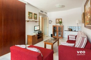 Een woonkamer met een rode bank, een glazen salontafel en een houten kast. Er staat een televisie op een dressoir en een deur die naar een andere kamer leidt.