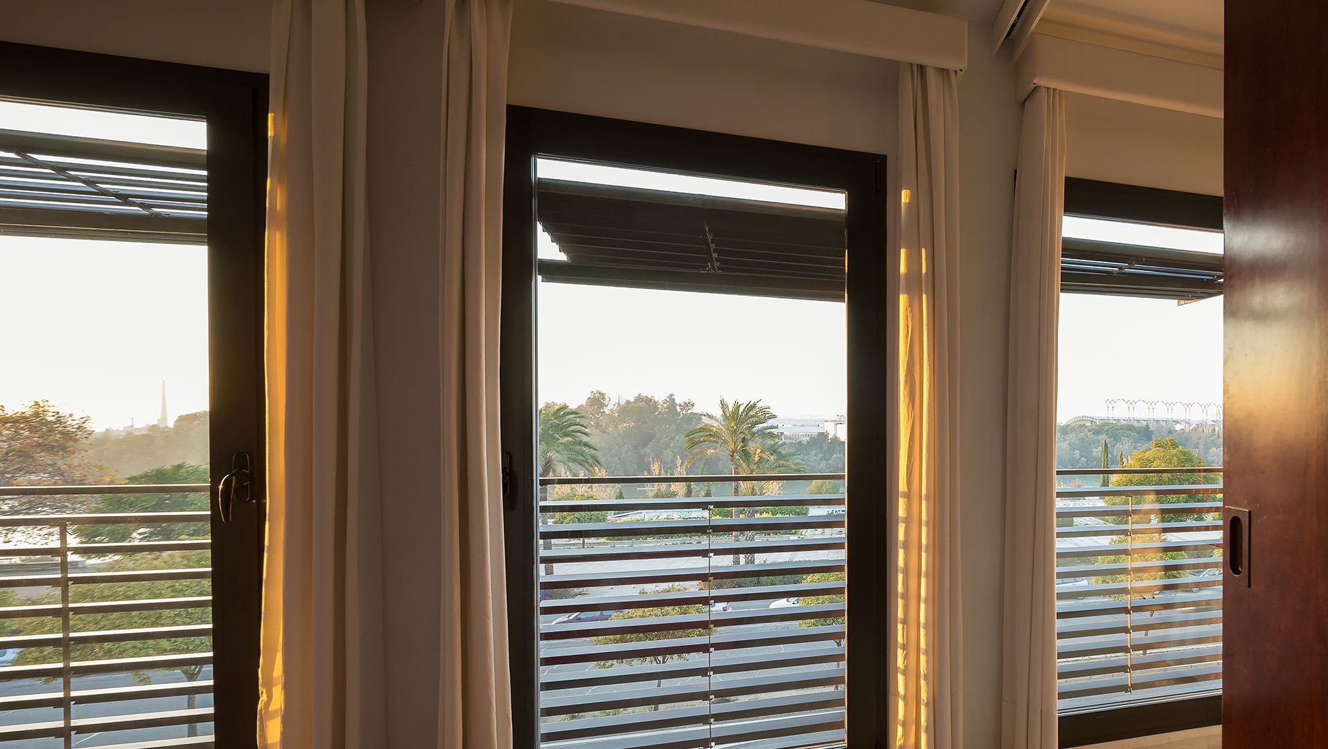 A room with large windows, white curtains, and a view of a cityscape.