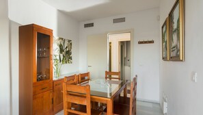 Uma área de refeições com um armário de madeira, uma mesa de vidro e cadeiras. Há um quadro na parede e uma porta que dá para outro cômodo.