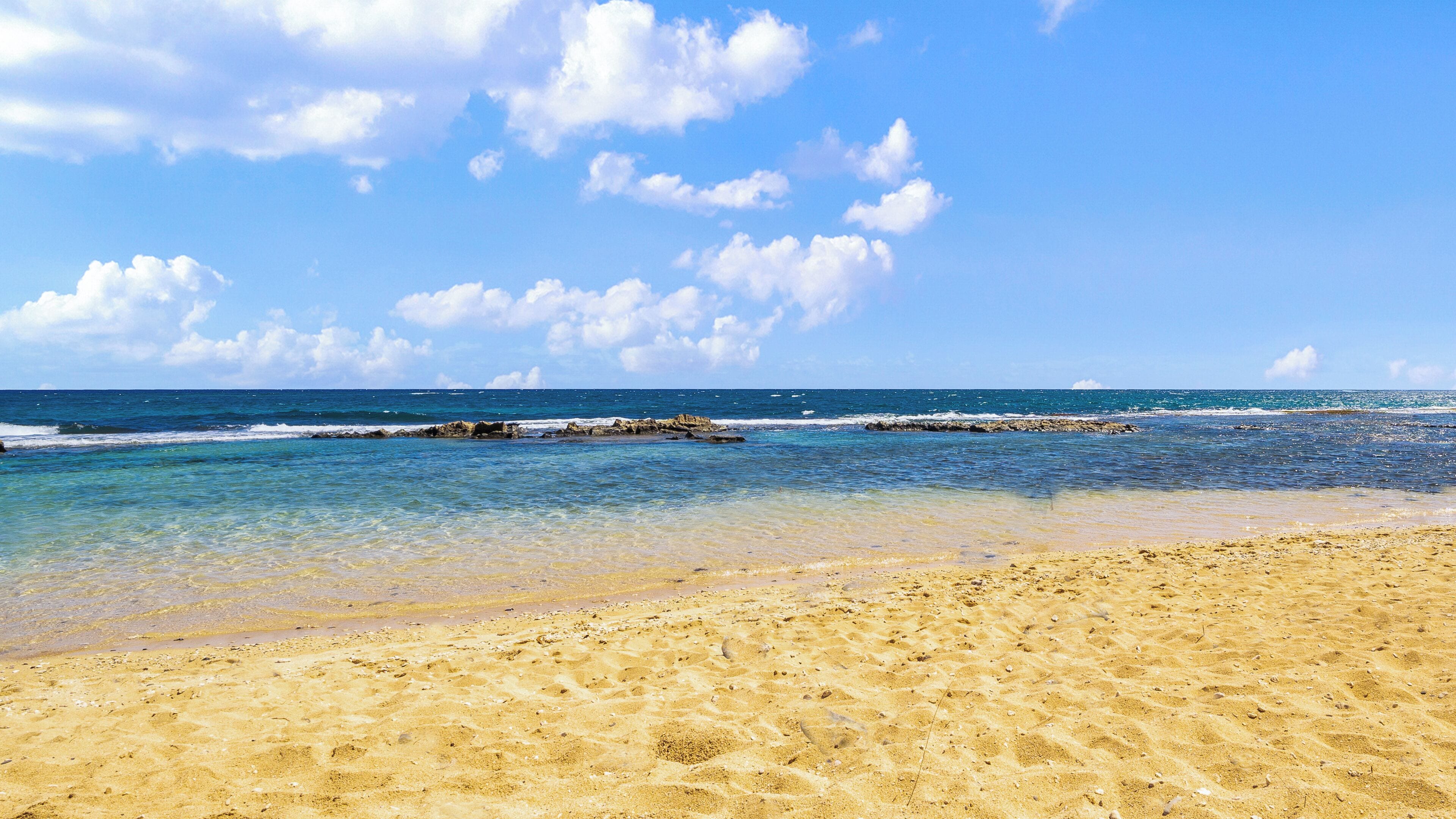 Una playa con aguas azul cristalinas, orilla arenosa y afloramientos rocosos.