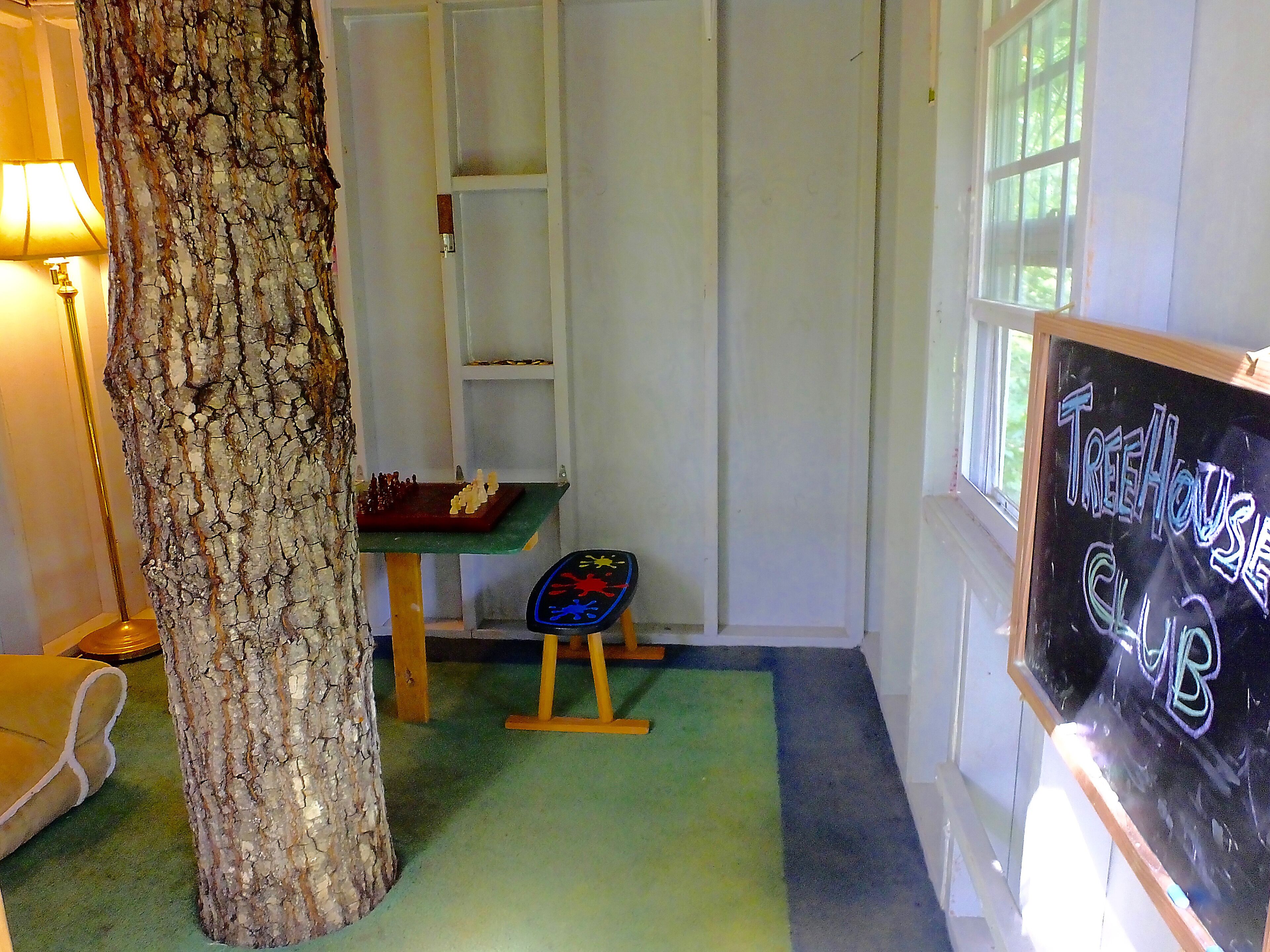 A room with a large tree trunk, a chessboard, a chalkboard with 'TreeHouse Club' written on it, and a window with natural light.