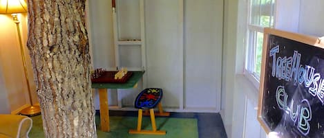 A room with a large tree trunk, a chessboard, a chalkboard with 'TreeHouse Club' written on it, and a window with natural light.