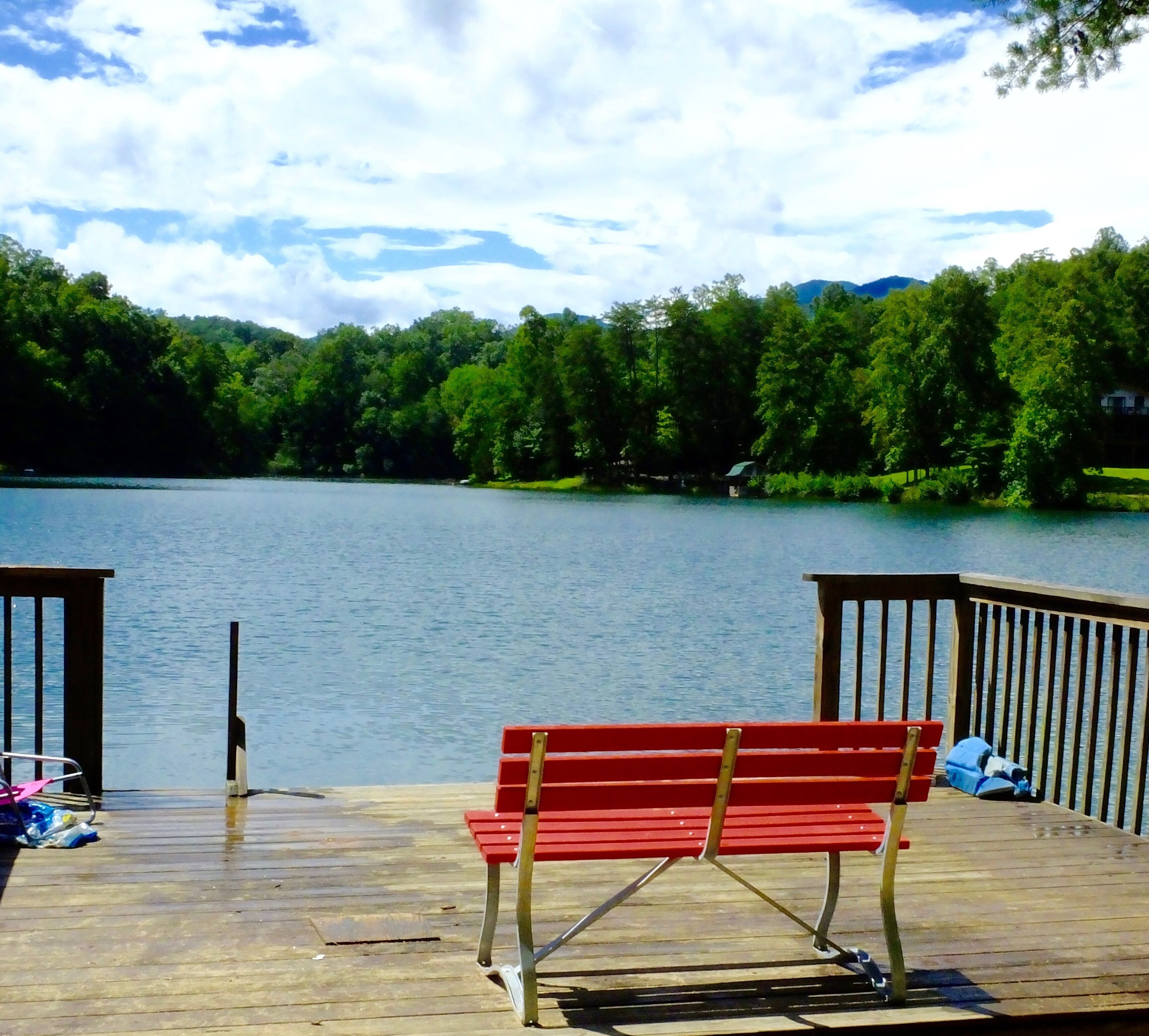 A wooden dock with a red bench overlooking a calm lake, surrounded by lush greenery and distant hills.