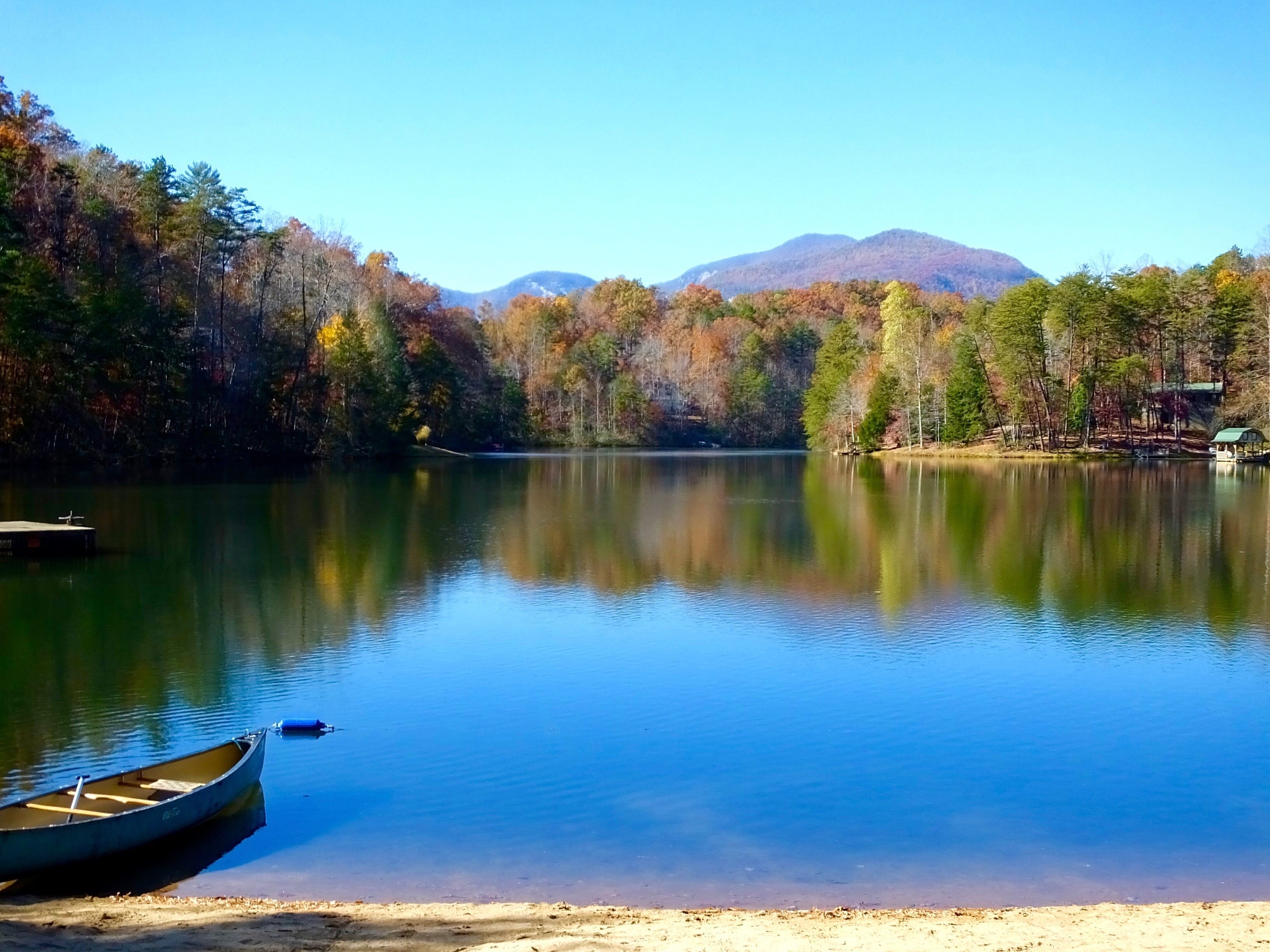 A calm lake with a dock and a small boat, surrounded by trees and mountains in the background.