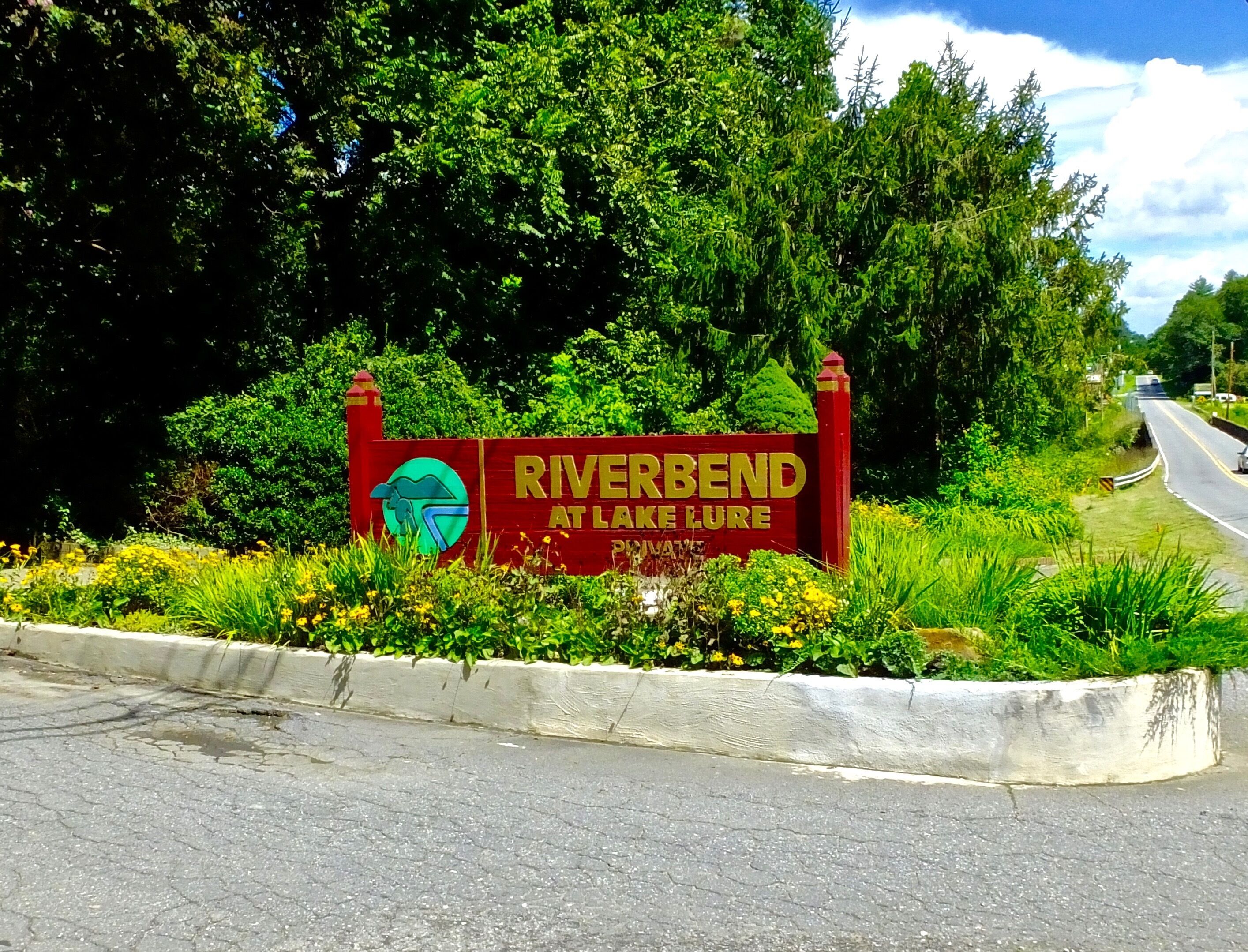 Un panneau rouge sur lequel on peut lire « RIVERBEND AT LAKE LURE », entouré de verdure et bordé par une route.