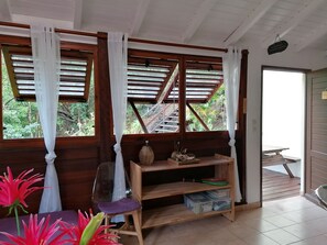 Una habitación con paredes de madera, una ventana con cortinas blancas, una estantería de madera con varios objetos, y una silla con una cubierta de plástico transparente.