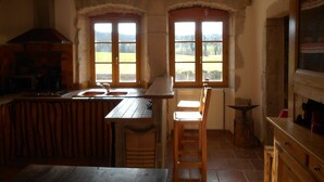 Cocina rústica con mesa de madera, un lavamanos y una estufa. Hay dos ventanas con vistas a un paisaje verde.