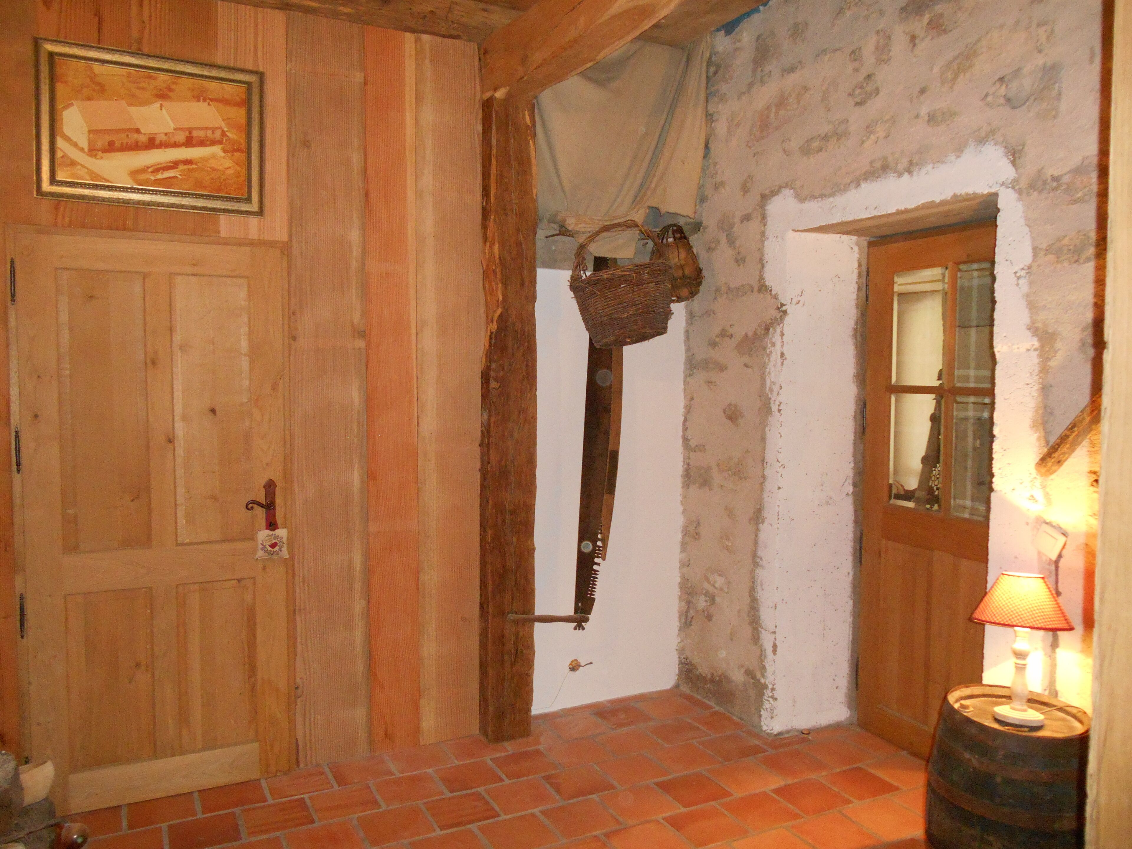 Une pièce rustique avec une porte en bois, un panier suspendu, une lampe posée sur un tonneau et un tableau encadré accroché au mur.