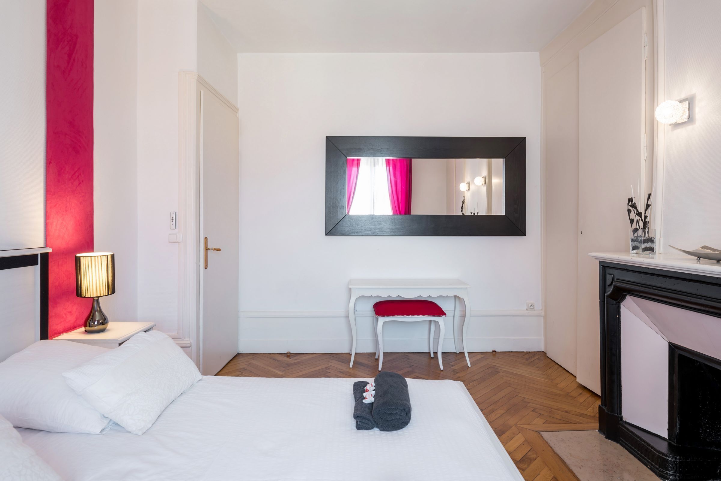 A hotel room with a bed, a nightstand with a lamp, a white bench, a mirror, and a fireplace.