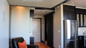 A modern kitchen with stainless steel appliances, a black leather sofa with an orange cushion, and a wooden floor.