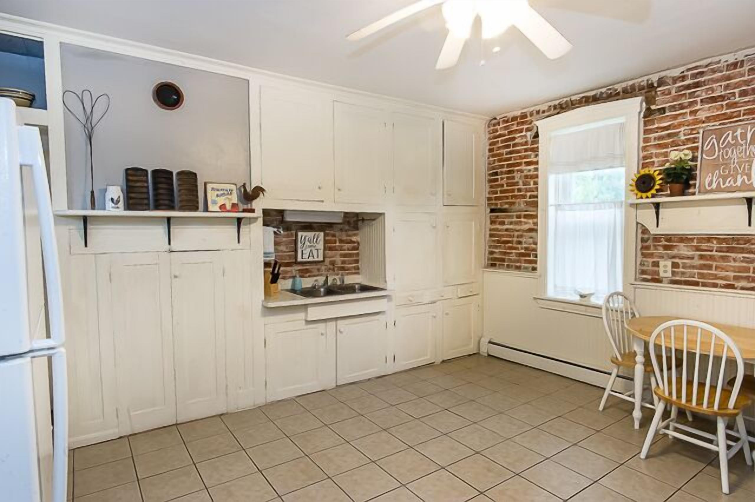 A kitchen with white cabinets, a brick wall, a window with a sunflower decoration, and a wooden table with chairs.