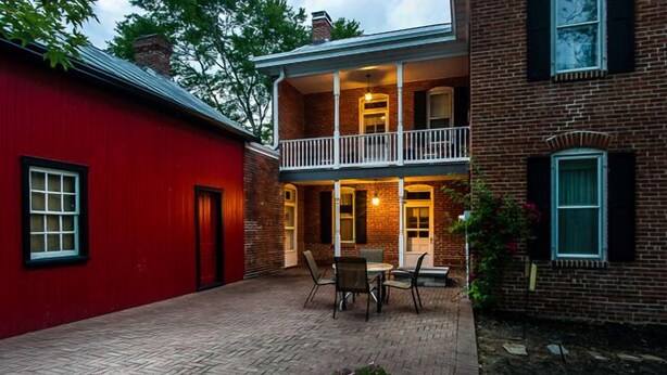 A two-story brick house with a balcony, a red exterior wall, and a brick patio with outdoor furniture.