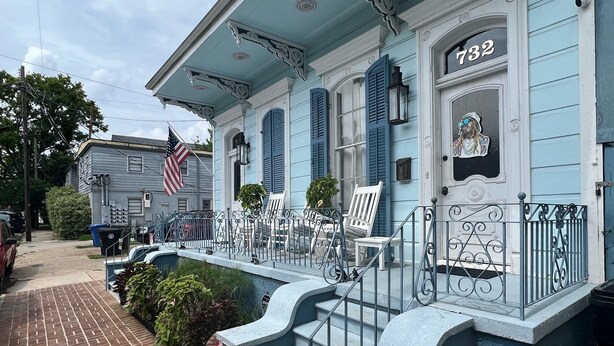 A blue house with a number 732 on the door, a brick pathway, and a small porch with two chairs and a potted plant.