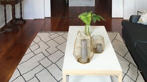 A living room with a white coffee table, a dark sofa, and a wooden side table with a lamp. A hallway leads to a kitchen area with a marble countertop and a water dispenser.