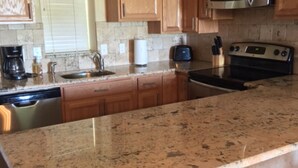 Una cocina con encimeras de granito, electrodomésticos de acero inoxidable, armarios de madera y un ventanal con persianas.