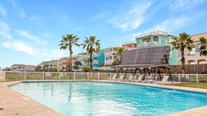 Una piscina con sillas de descanso y una estructura de madera, rodeada de edificios coloridos y palmeras.