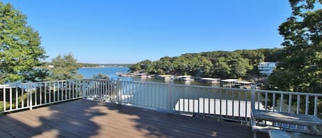 A wooden deck with a white railing overlooking a marina with boats and a forested shoreline.