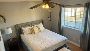 Une chambre à coucher avec un lit, une lampe de chevet, un ventilateur de plafond et une fenêtre avec des rideaux.