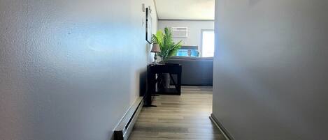 Un pasillo con suelo de madera, paredes blancas y un techo con una rejilla de ventilación. Se ve una planta sobre una mesa al fondo.