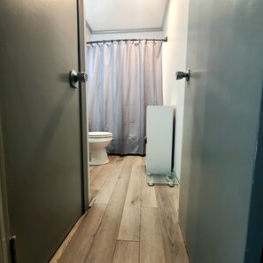 Ein schmaler Flur mit Holzboden führt zu einem Badezimmer. Im Badezimmer sind eine Toilette und ein weißes Schränkchen zu sehen.