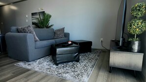A modern living room with a grey sofa, a small black ottoman, a patterned rug, and a minimalist side table.