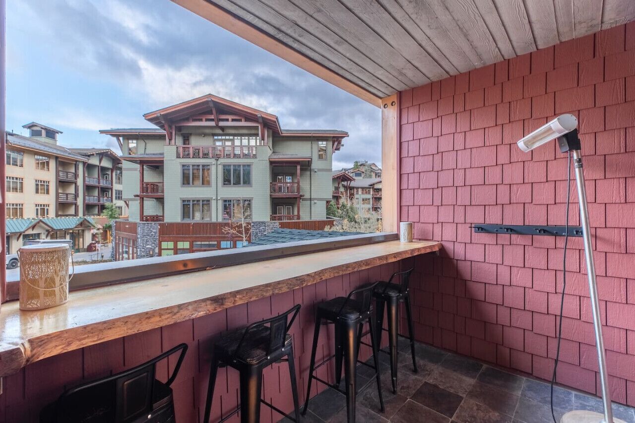 A bar area with a wooden counter, black bar stools, and a view of a multi-story building with balconies.