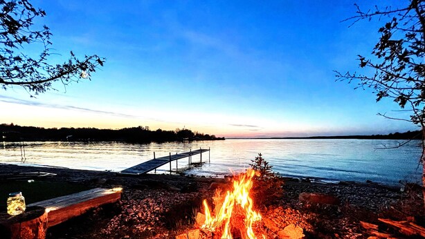 A lakeside scene with a dock extending into calm water, a fire burning on the shore, and a clear sky.