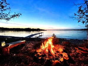 Maisema järven rannalla: laiturinokka ulottuu tyynen veden ylle, rannalla palaa nuotio ja taivas on kirkas.