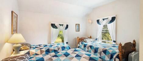 A bedroom with two beds, each with a geometric-patterned bedspread, a bedside table with a lamp, and a fan.