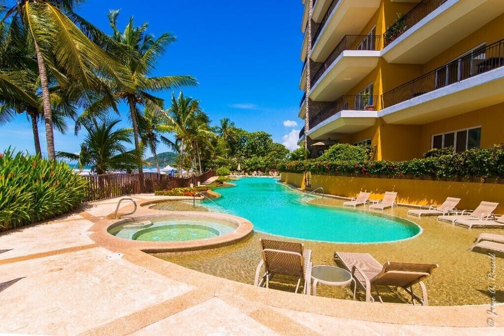 A hotel pool area with multiple lounge chairs and a hot tub, surrounded by palm trees and a clear blue sky.