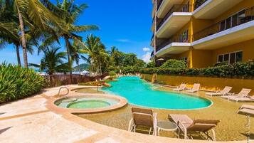 A hotel pool area with multiple lounge chairs and a hot tub, surrounded by palm trees and a clear blue sky.