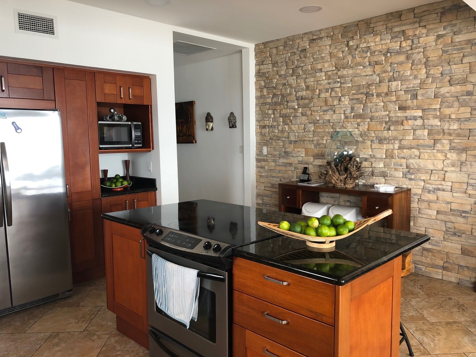 A kitchen with a stone wall, wooden cabinets, a stainless steel refrigerator, a microwave, and a stove.