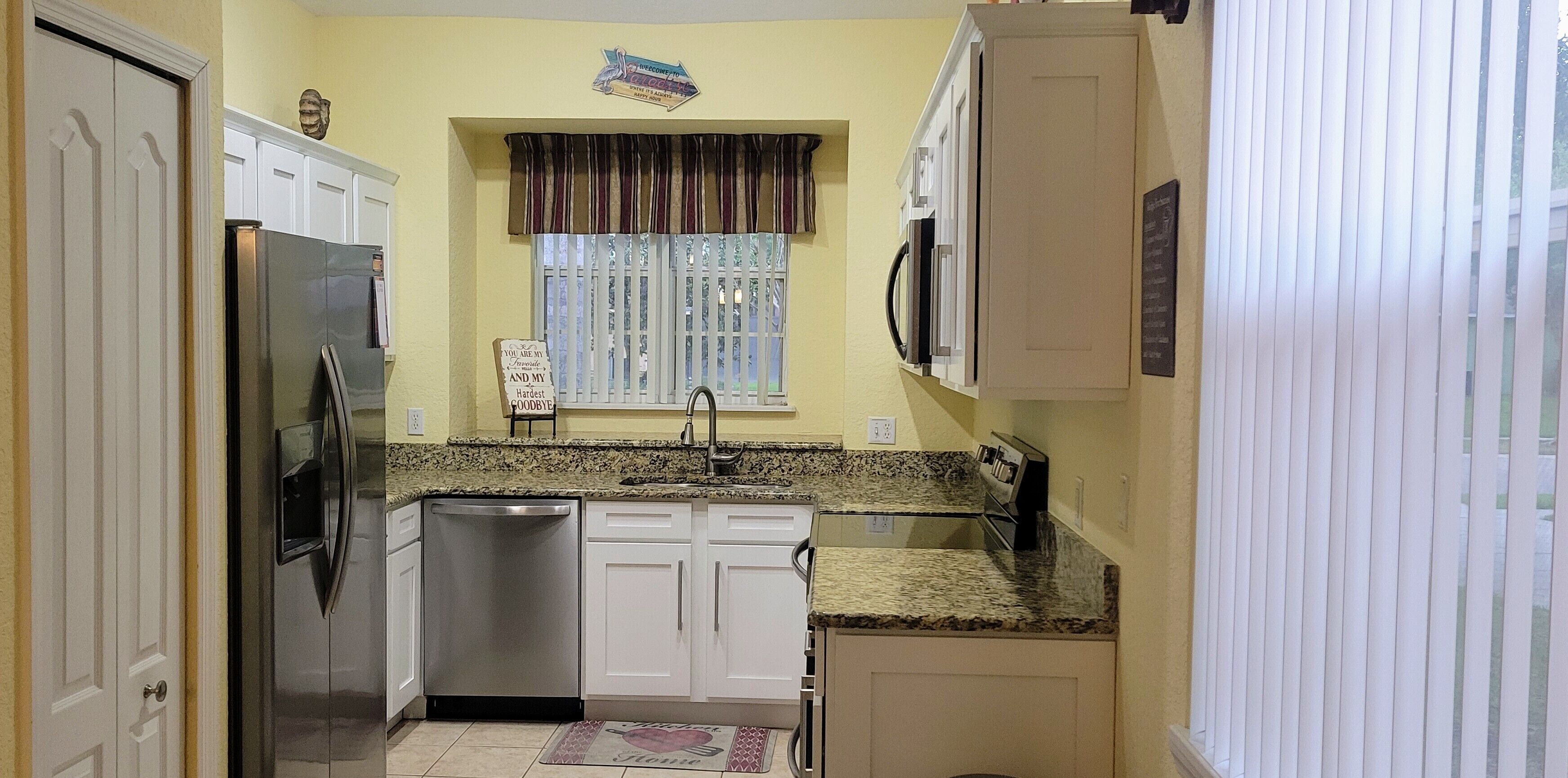A compact kitchen with white cabinets, a stainless steel refrigerator, a dishwasher, and a window with a striped valance.