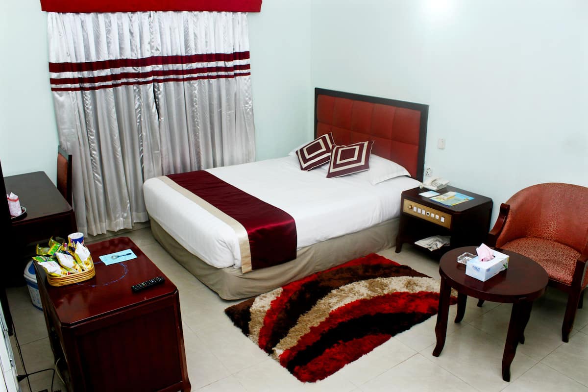 A hotel room with a bed, a nightstand, a chair, a small table, a remote control, and a red and beige rug.