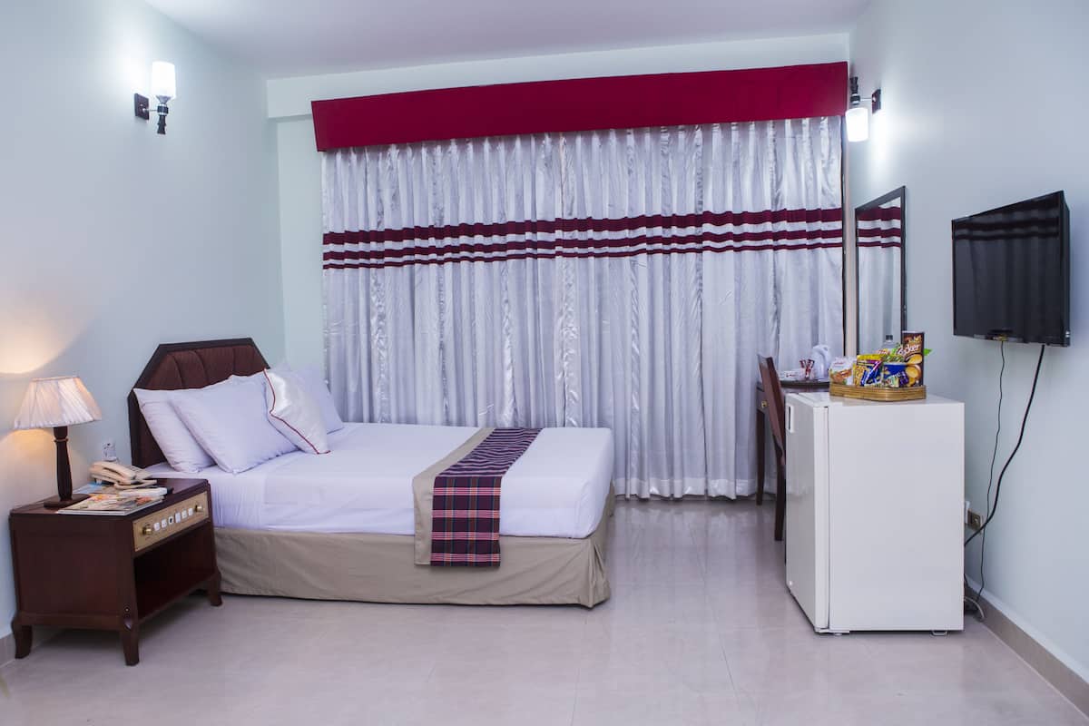 A hotel room with a bed, a nightstand with a lamp, a desk with a television, and a refrigerator.