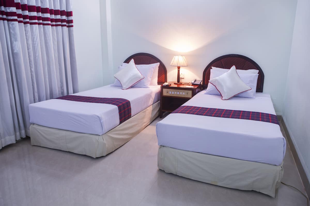 A hotel room with two single beds, each with a pillow and a blanket. There is a nightstand with a lamp between the beds.