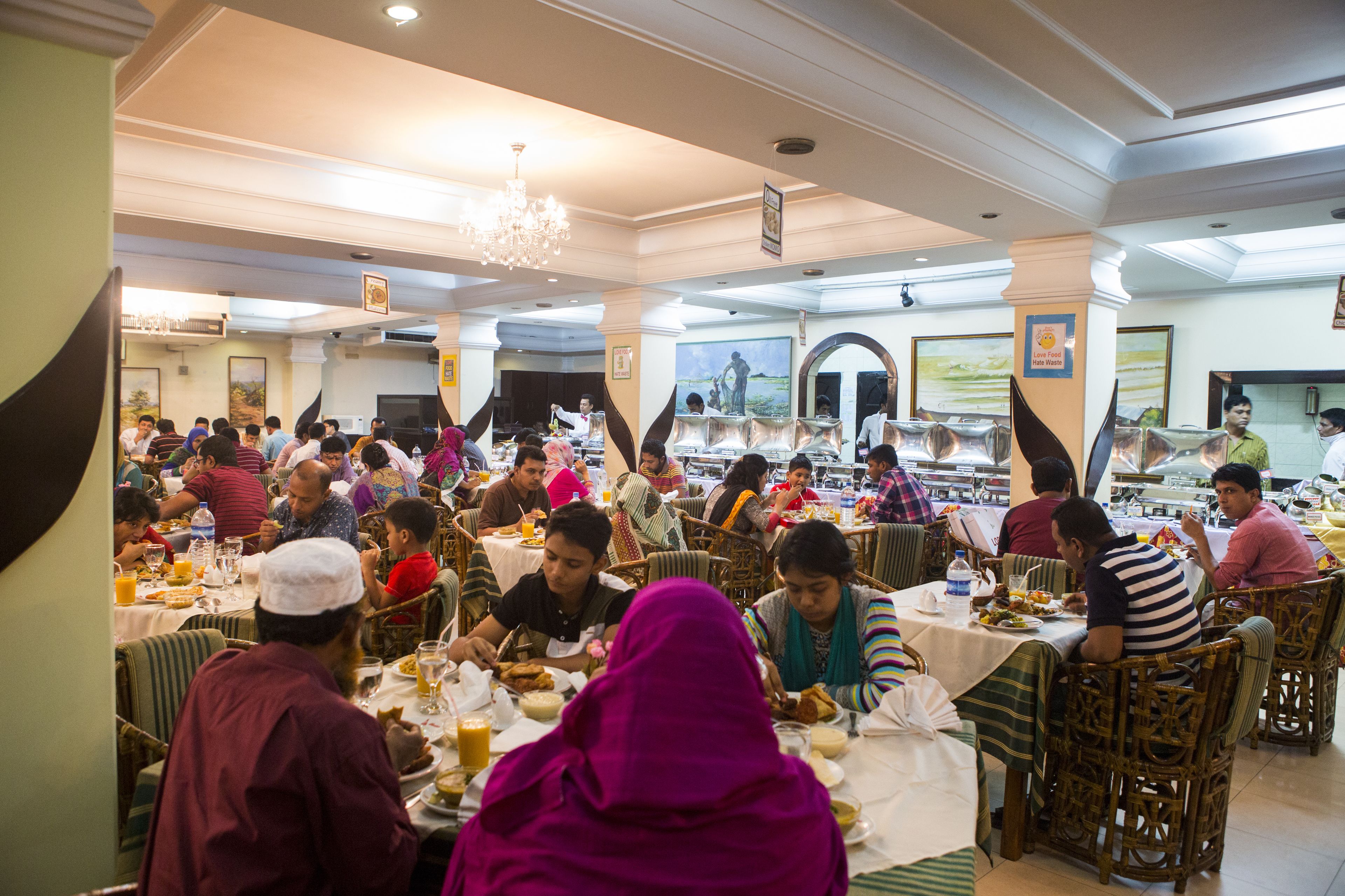 A crowded dining area with tables and chairs, people eating, and a chandelier.