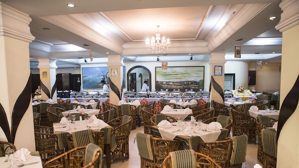 A restaurant with wicker chairs and tables, a chandelier, and a large painting on the wall. - Meridian Hotel (Chittagong)