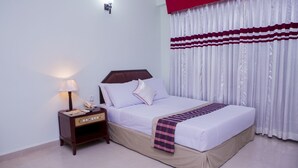 A neatly made bed with white linens and a plaid blanket, a bedside table with a lamp, and a window with white curtains. - Meridian Hotel (Chittagong)