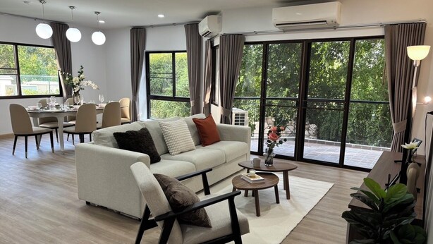 A modern living room with a sofa, armchair, dining table, and chairs. There are large windows with curtains, a potted plant, and an air conditioning unit. - Seastar Hotel And Service Apartment (Ban Chang)