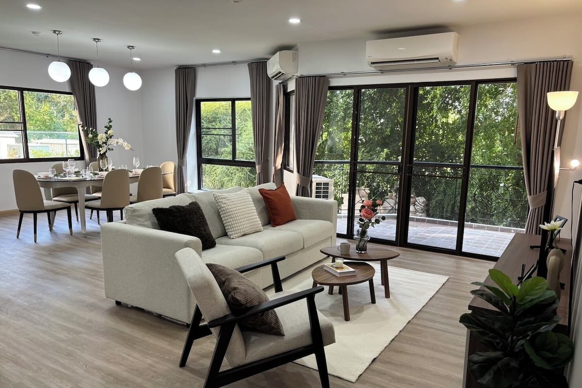 A modern living room with a sofa, armchair, dining table, and chairs. There are large windows with curtains, a potted plant, and an air conditioning unit.
