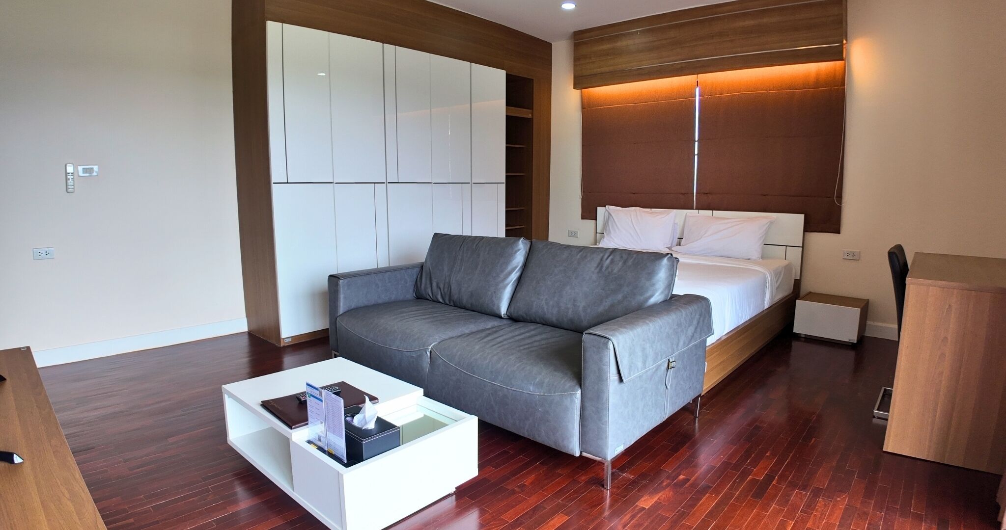 Una habitación de hotel moderna con un sofá gris, una mesa de centro blanca, una cama con ropa de cama blanca y un armario de madera.