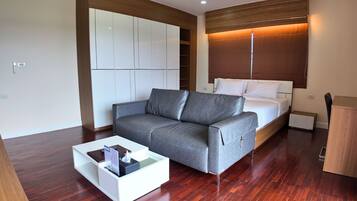 Una habitación de hotel moderna con un sofá gris, una mesa de centro blanca, una cama con ropa de cama blanca y un armario de madera.