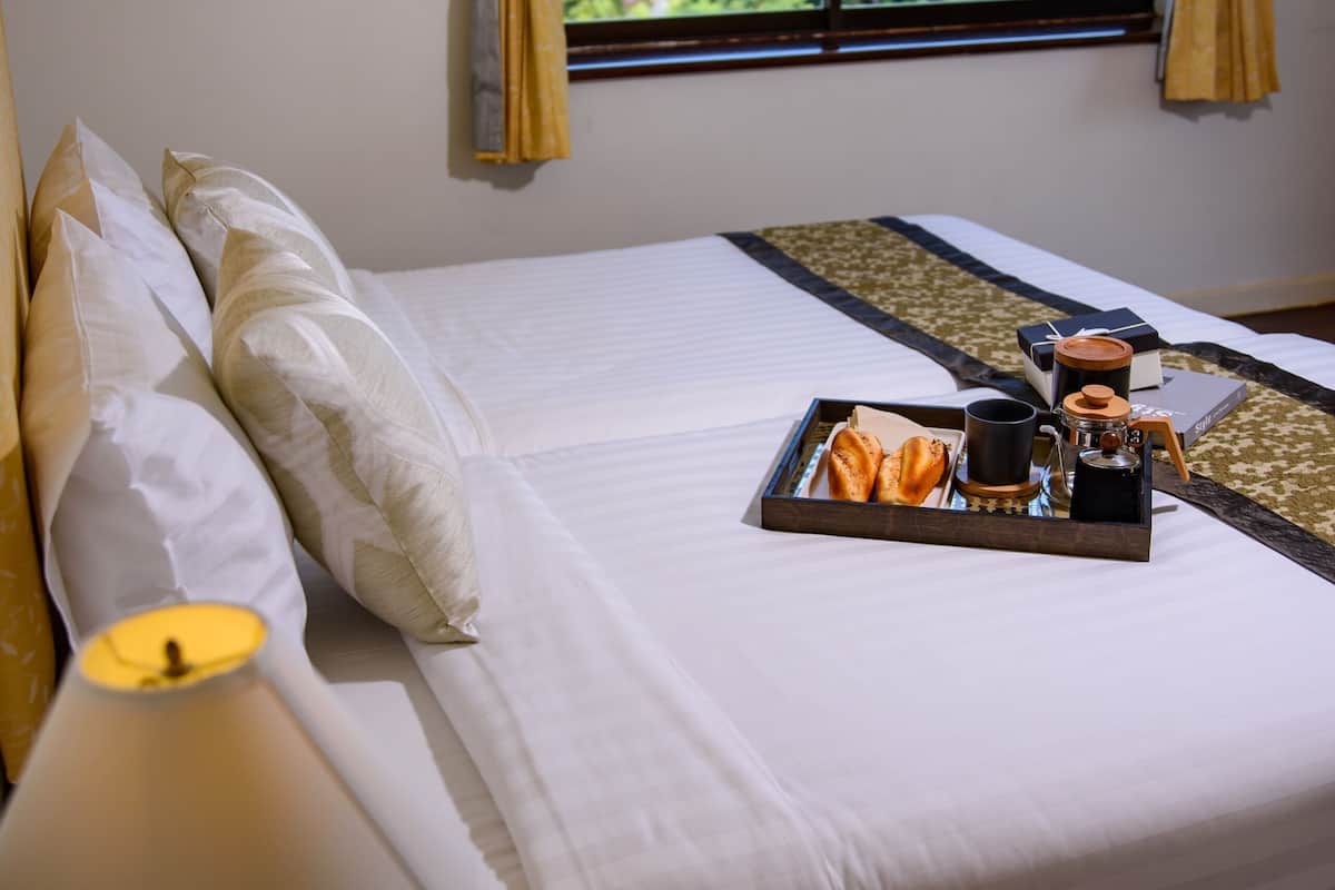 A neatly made bed with a tray of breakfast items, a bedside lamp, and a window with curtains.
