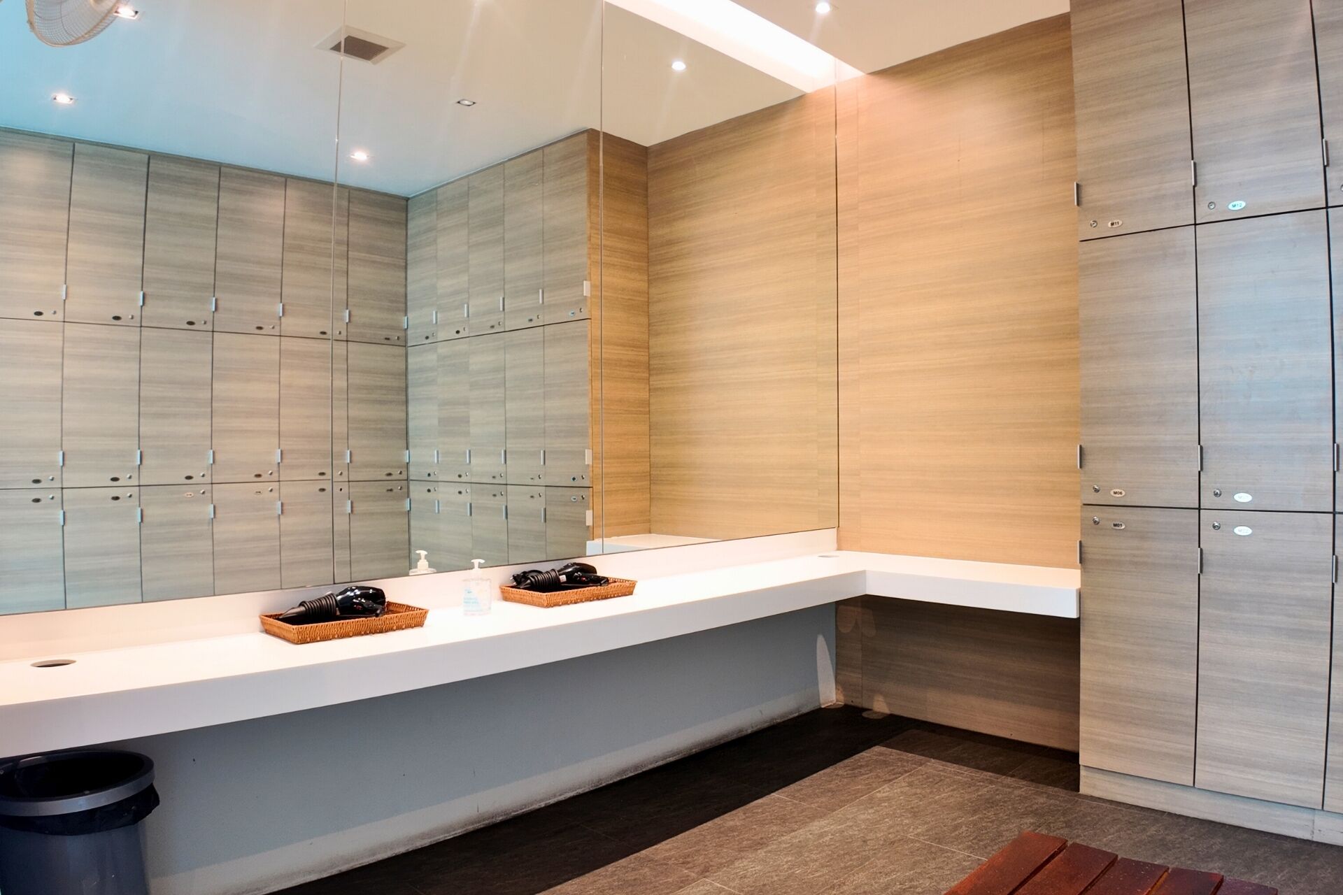 A modern bathroom with a large mirror, wooden lockers, a white vanity, and a trash bin.