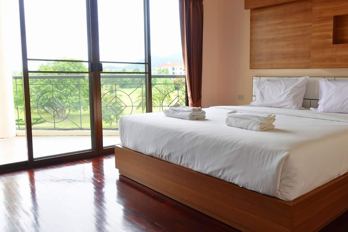A modern bedroom with a large bed, wooden headboard, and a view of greenery through large windows.