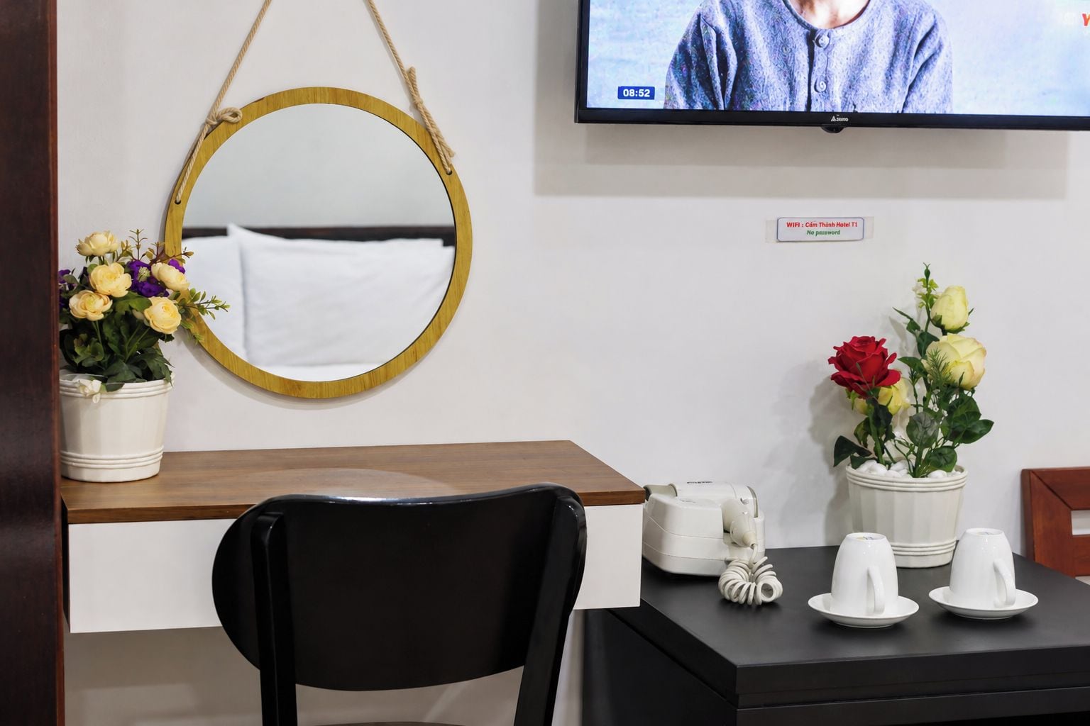A hotel room with a wooden desk, a black chair, a round mirror, and two potted plants with flowers.