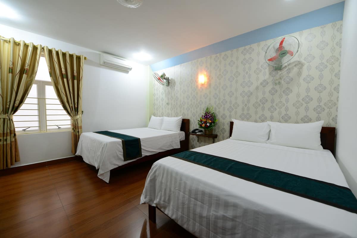 Een hotelkamer met twee eenpersoonsbedden, een houten vloer, een raam met gordijnen, een wandventilator en een tafeltje met een bloevas.