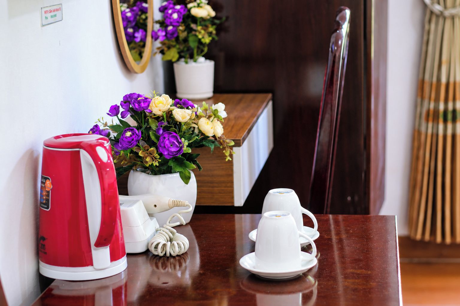 A hotel room with a wooden table, a red electric kettle, a white coffee maker, a white teapot, a white cup, and a vase with purple and white flowers.