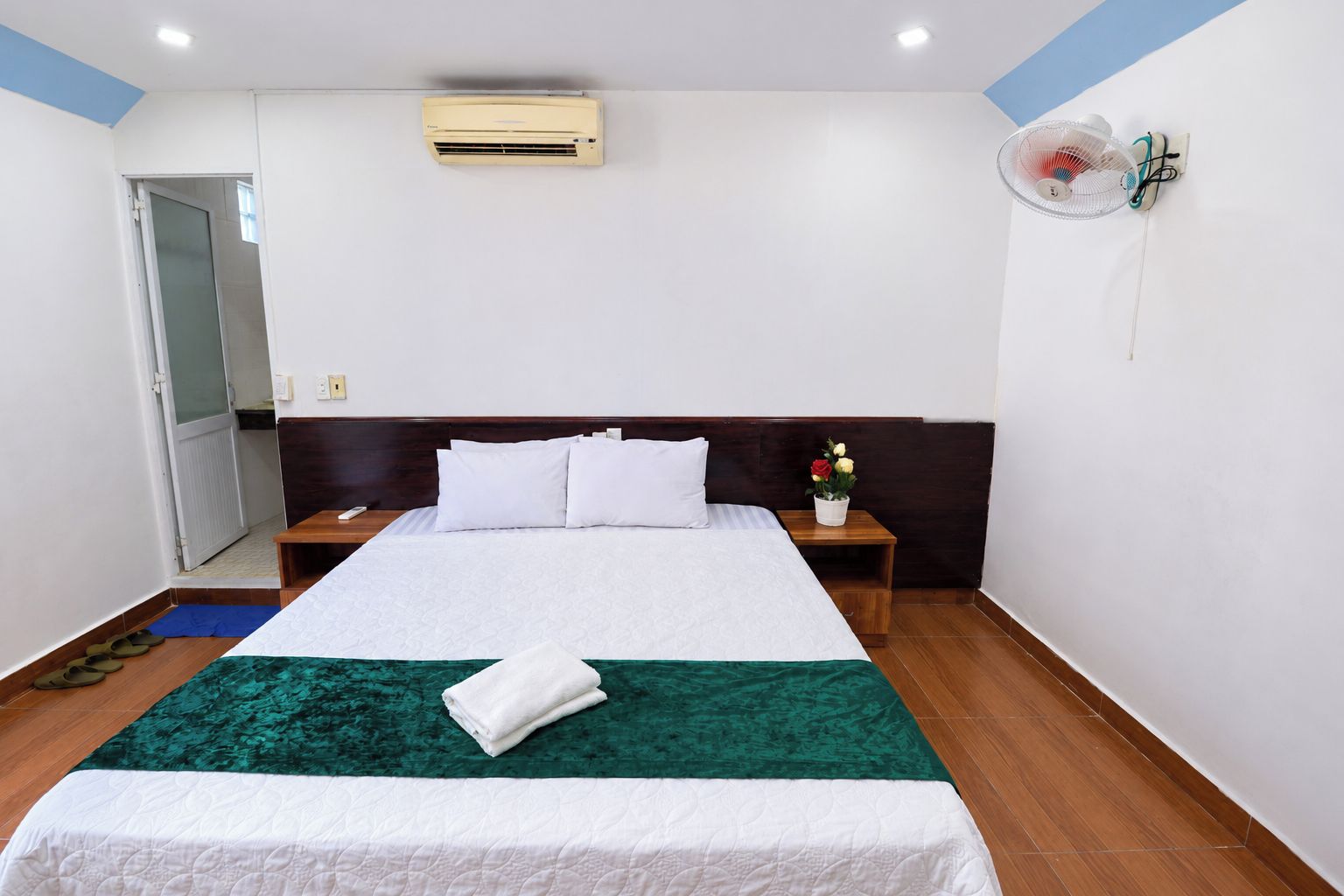 A hotel room with a bed, a bedside table, a nightstand, a wall-mounted air conditioner, and a wall fan.